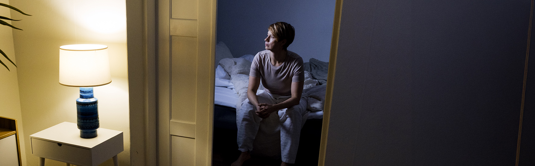 A person sits on a bed in a dim room, illuminated by a table lamp.