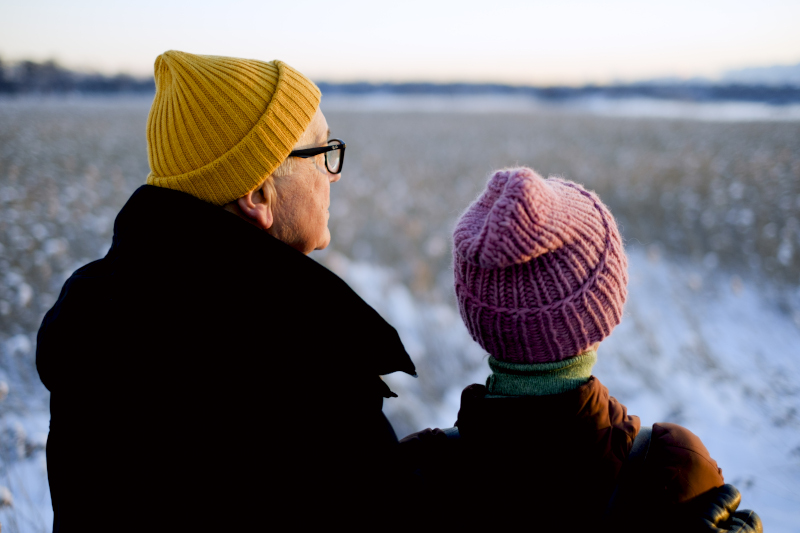Ikääntyneet mies ja nainen seisovat selin kameraan ja katsovat talvista maisemaan, värikkäät pipot päässään.
