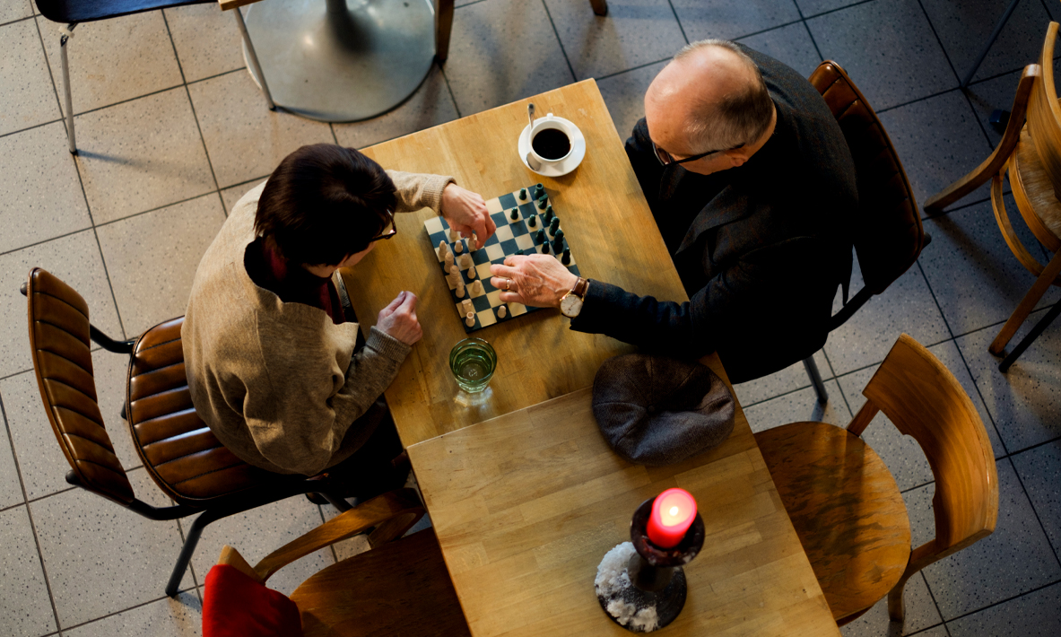 Ikääntynyt nainen ja mies pelaavat shakkia pöydän ääressä.