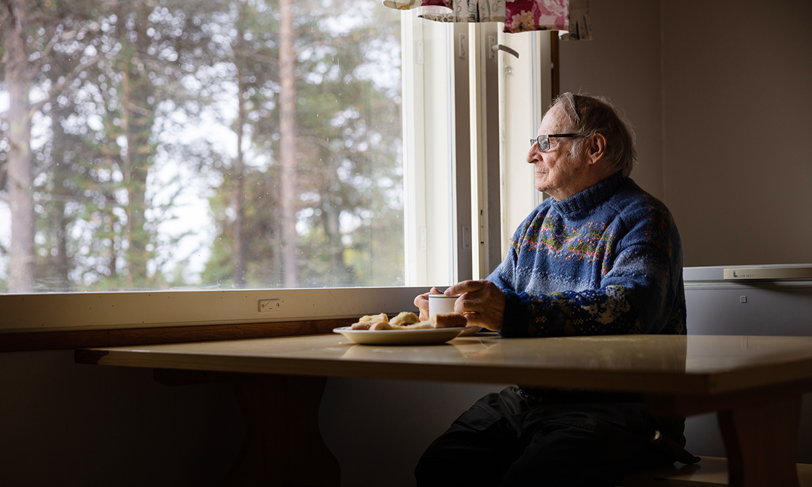 En gammal man i tröja sitter vid ett bord, dricker kaffe och tittar ut genom fönstret. 